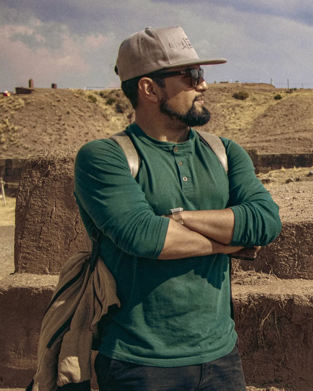Juan André Hidalgo Ugalde con los brazos cruzados, gorra, lentes de sol y mochila, junto a muros de piedra en un paisaje de altiplano
